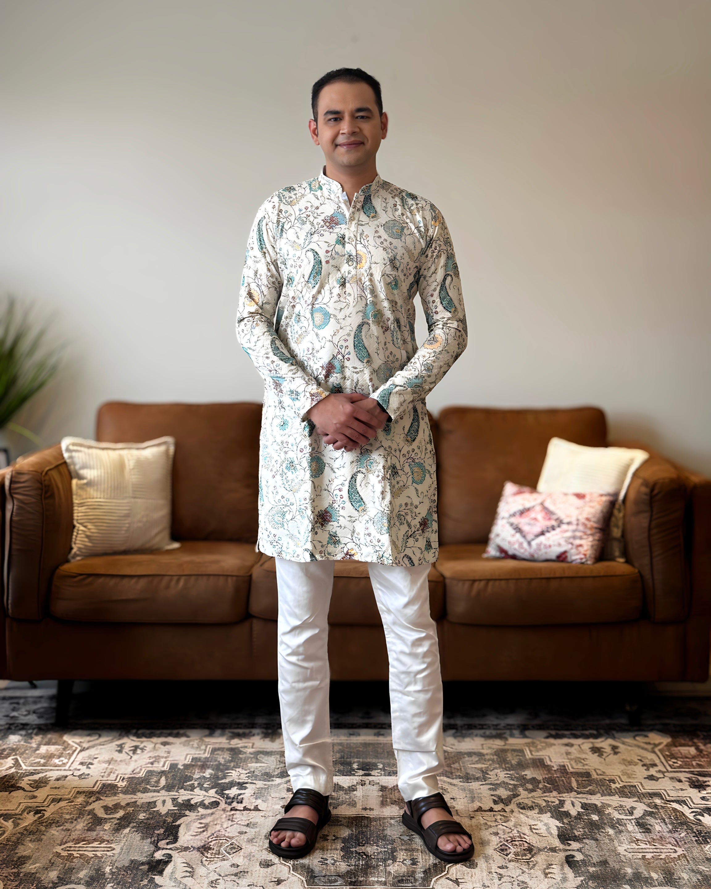 Man wearing a patterned kurta standing in a living room with a brown sofa and decorative pillows.