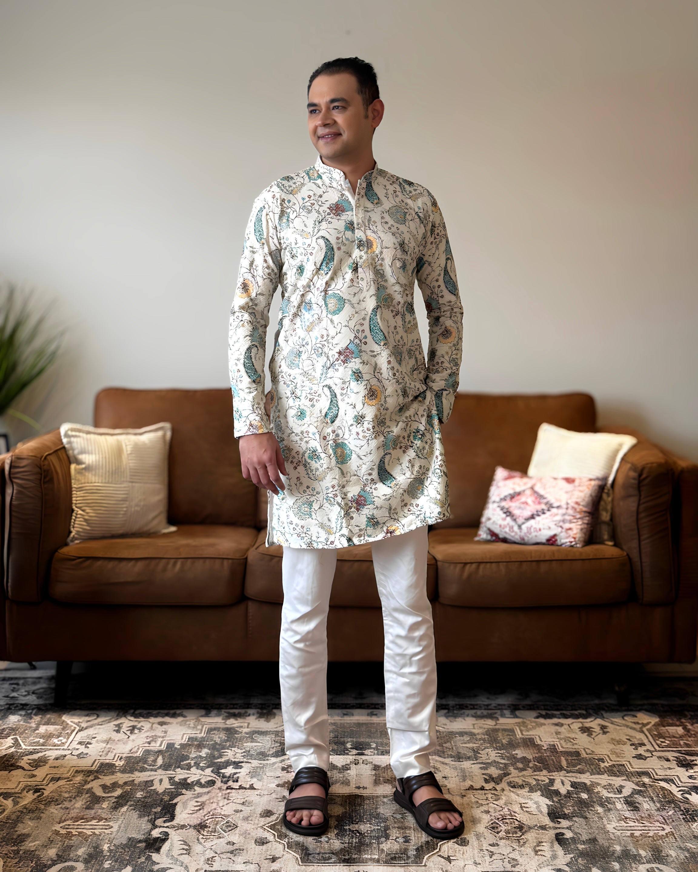 Man wearing a floral kurta standing in a living room with a brown sofa and patterned rug.