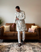 Man wearing a patterned kurta standing in a living room with a brown sofa and decorative pillows.