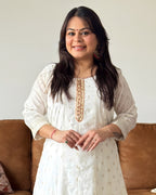 Woman in a white traditional outfit with gold embroidery sitting on a brown couch.