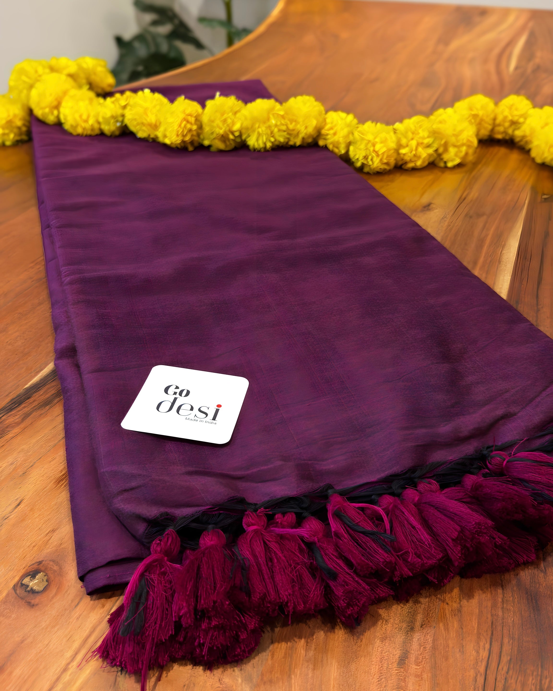 Purple scarf with yellow pom-poms on a wooden surface, featuring the 'Go desi' brand.