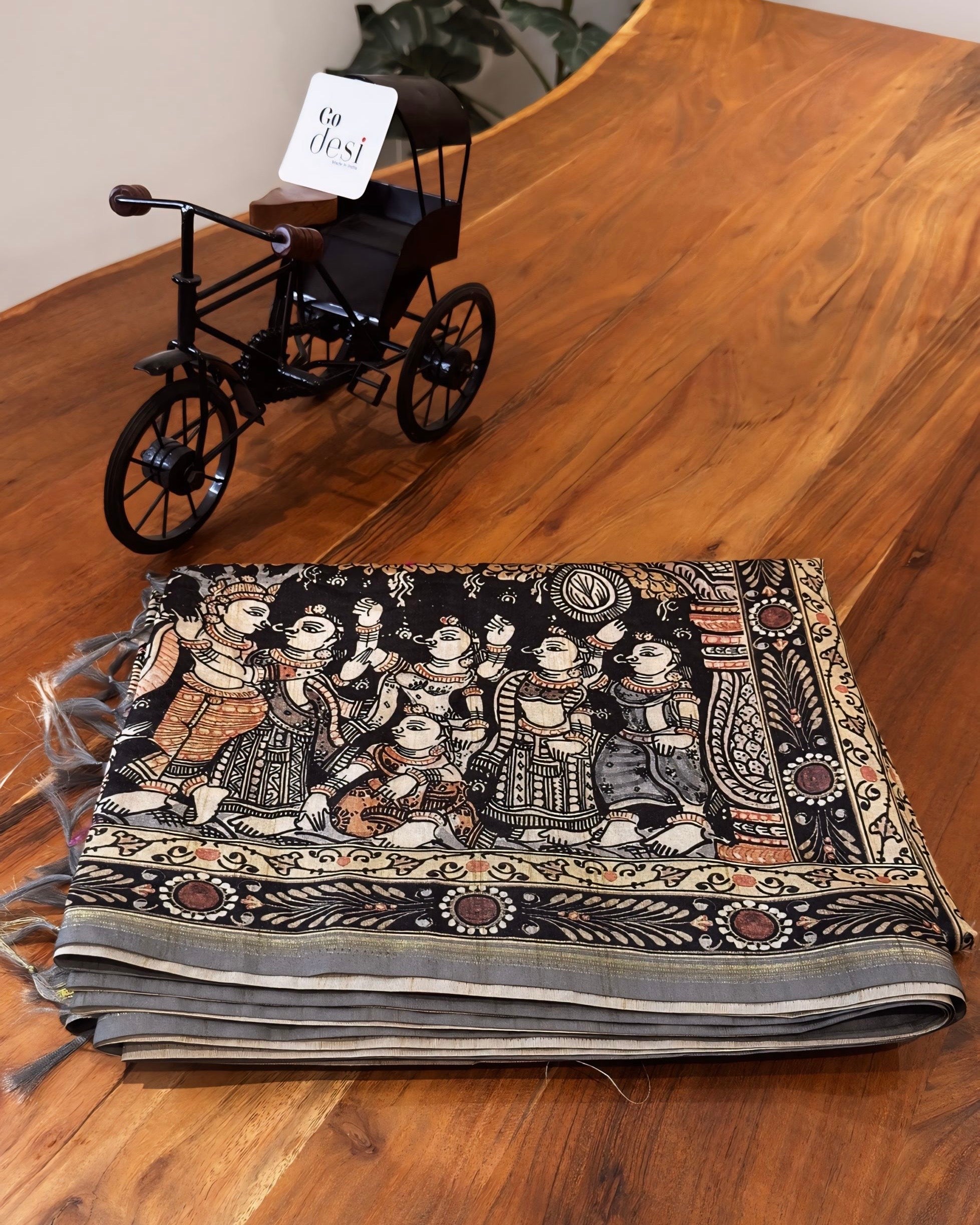 Decorative scarf with intricate patterns on a wooden surface, with a small model tricycle beside it.