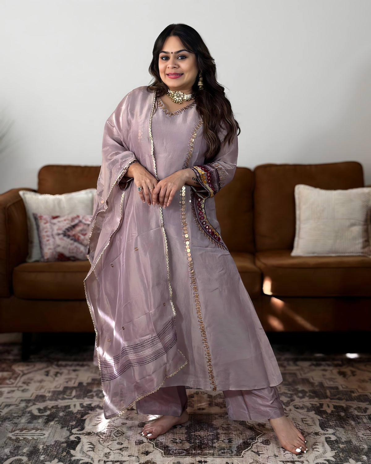 A woman modeling a light lavender-purple traditional South Asian outfit with gold embroidery, standing in front of a brown sofa and patterned rug.