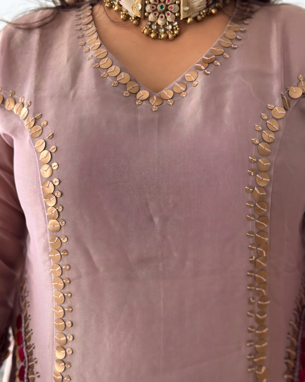 A close-up of a dusty pink or mauve-colored traditional garment with ornate gold coin embroidery along the V-neckline and down the front panels. A chunky gold-toned statement necklace is also visible at the neck.