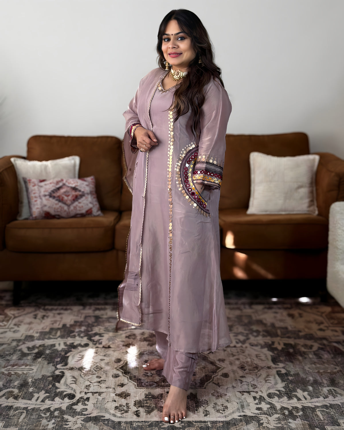 A woman stands barefoot in a living room wearing a light dusty pink traditional outfit with gold embroidery and a matching long coat. The background features a brown sofa with decorative pillows and a patterned rug.
