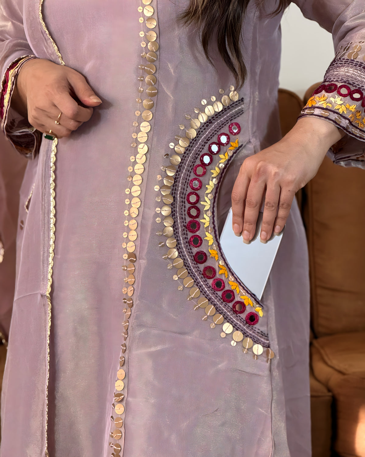 A close-up of a person wearing a lavender embroidered tunic with gold coin embellishments and a decorative multicolor mirror-work panel on the front. The person's hands are visible holding a phone near the embroidery.