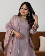 A woman wearing a lavender-purple traditional embroidered kurta set with gold mirror work and metallic trim, standing in front of a brown sofa.