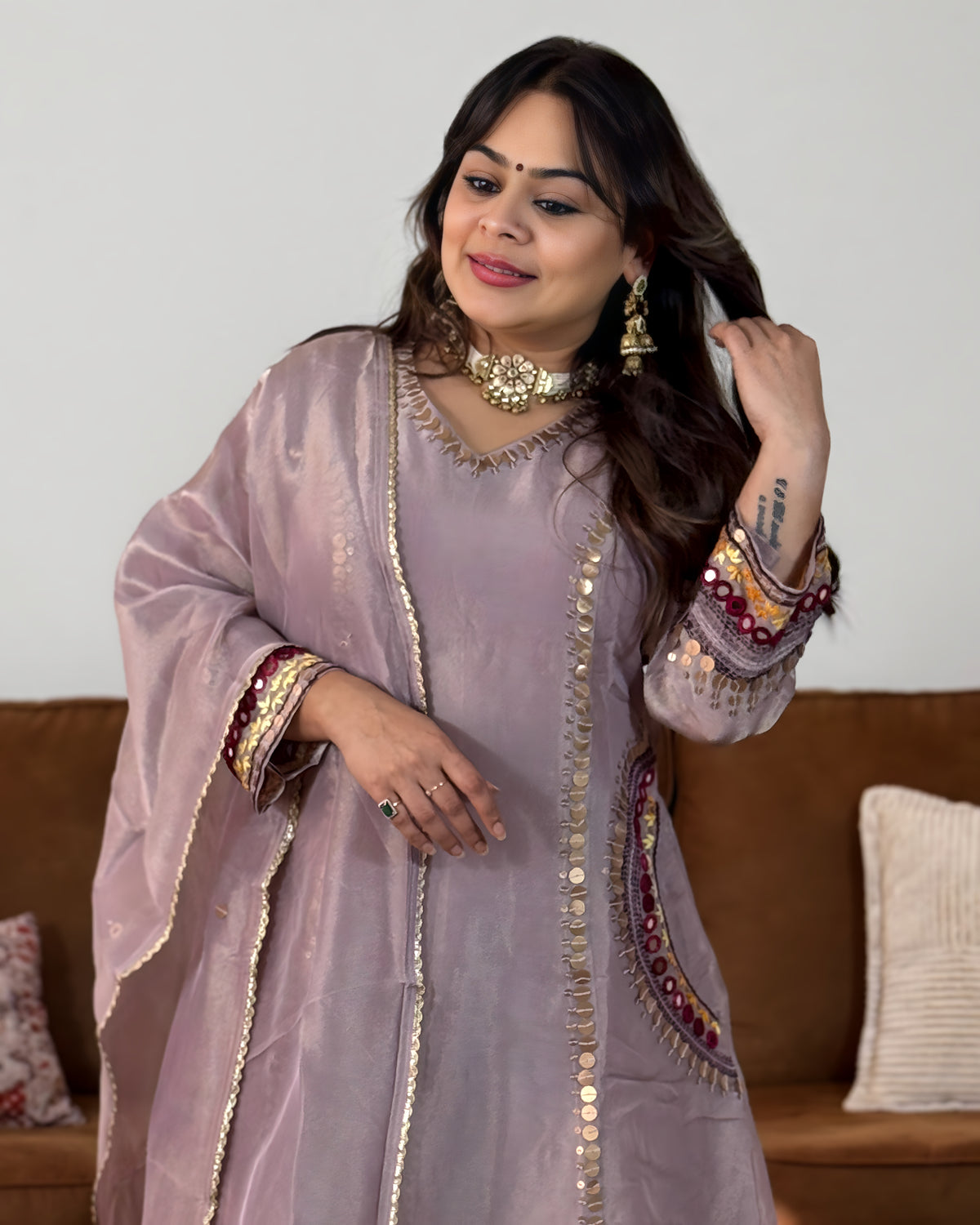 A woman wearing a lavender-purple traditional embroidered kurta set with gold mirror work and metallic trim, standing in front of a brown sofa.