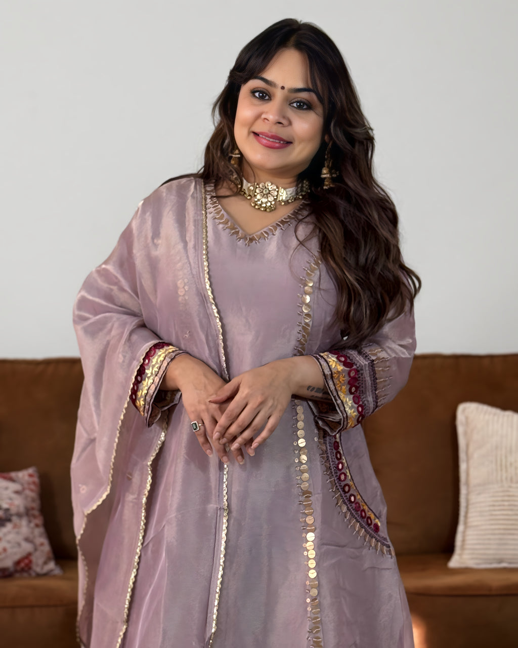 A woman wearing a lavender traditional outfit with gold coin embroidery and colorful patterned cuffs, standing in front of a brown sofa.