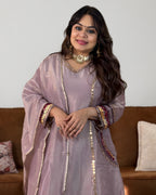 A woman wearing a lavender traditional outfit with gold coin embroidery and colorful patterned cuffs, standing in front of a brown sofa.