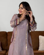 A woman wearing a lavender embroidered kurta with gold coin embellishments and red accents, standing in front of a brown sofa.