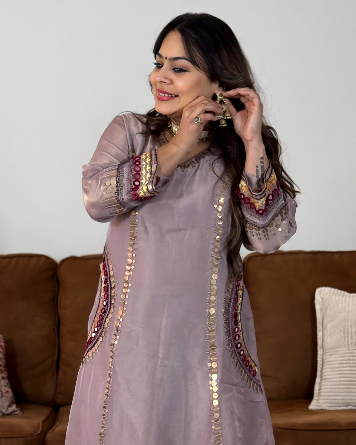 A woman wearing a lavender embroidered kurta with gold coin embellishments and red accents, standing in front of a brown sofa.