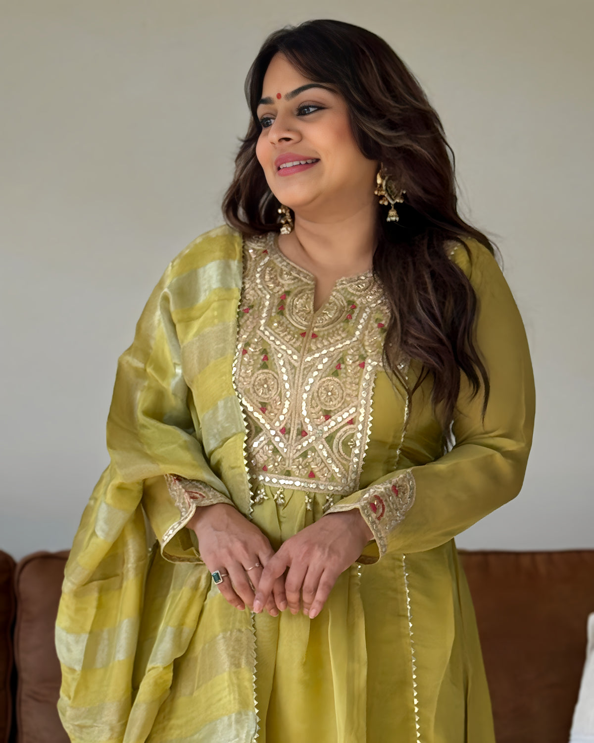 A woman wearing a traditional Indian outfit in olive green with intricate gold embroidery on the chest and cuffs, paired with a matching sheer dupatta, standing indoors.