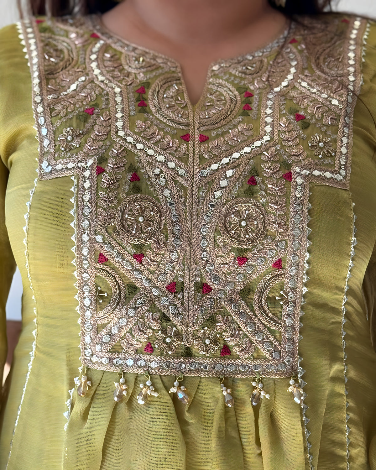 Close-up of a traditional embroidered kurta in olive green, featuring intricate gold and silver mirror work, beadwork, and small red floral accents on the yoke.