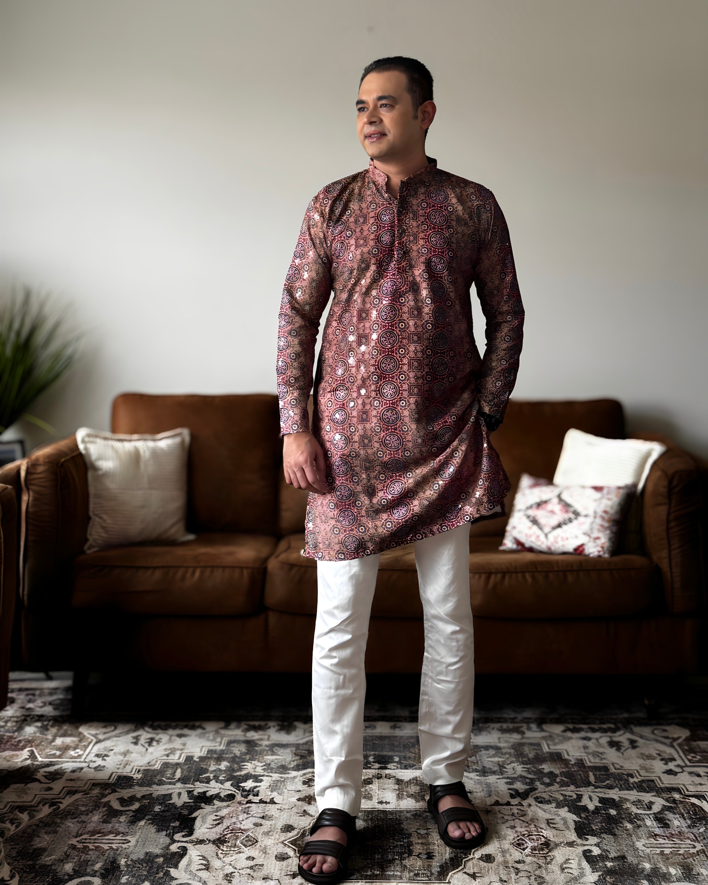 Man wearing a patterned kurta with white pants standing in a living room.