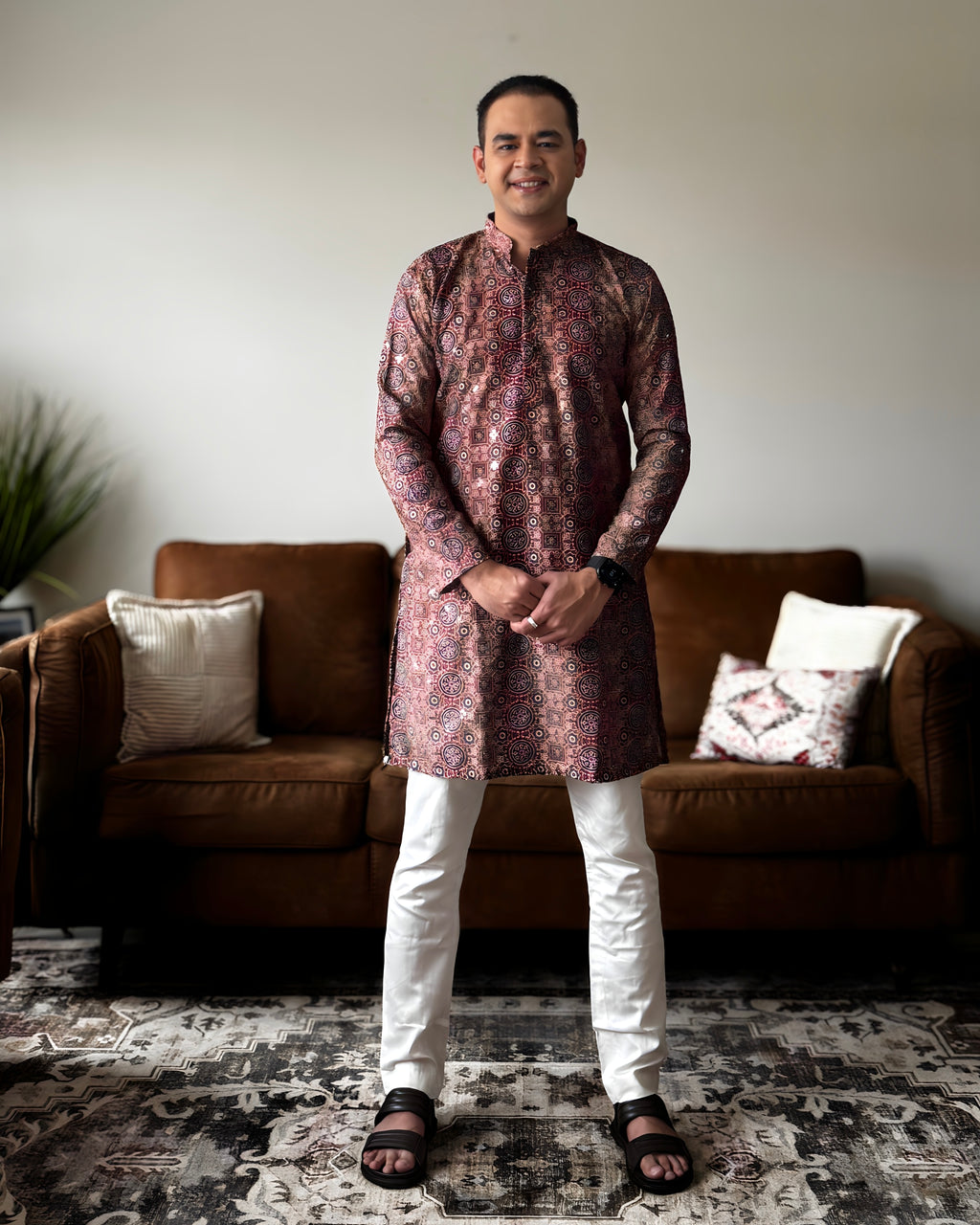 Man wearing a patterned kurta standing in a living room with a brown sofa and decorative pillows.