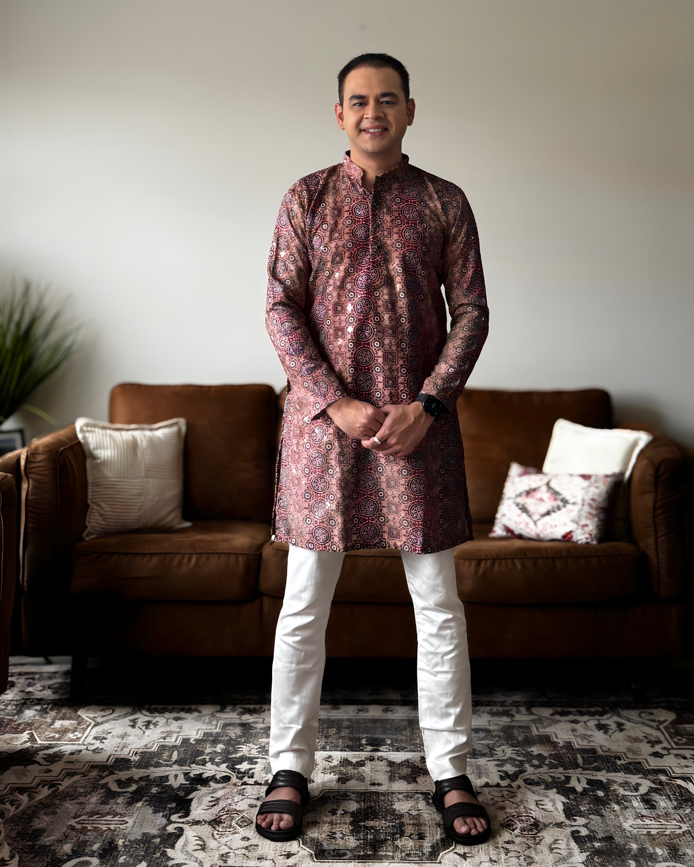 Man wearing a patterned kurta standing in a living room with a brown sofa and decorative pillows.