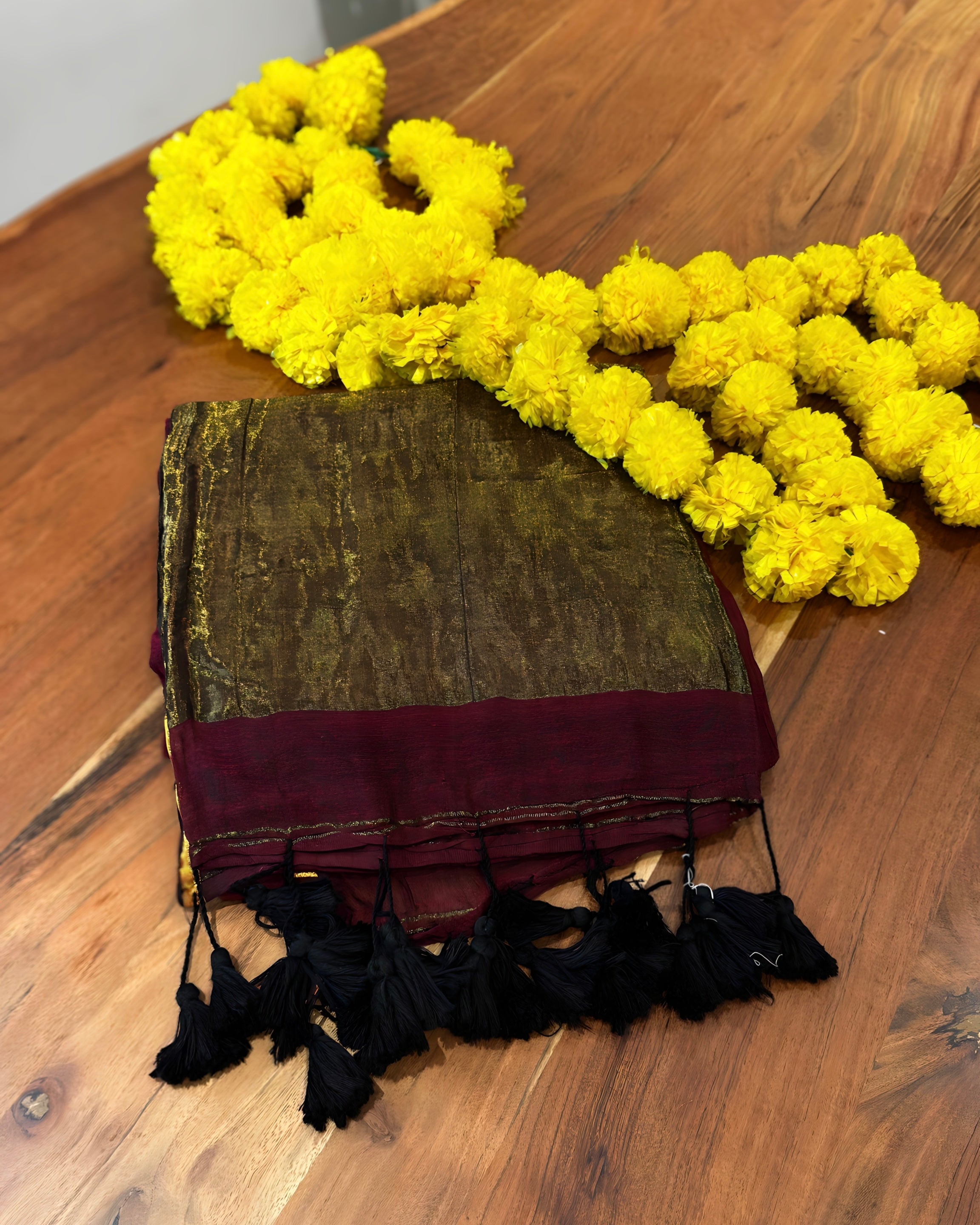 Green and maroon fabric with black tassels on a wooden surface, accompanied by yellow flowers.