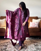 Woman in a purple traditional outfit holding a matching dupatta in a living room.
