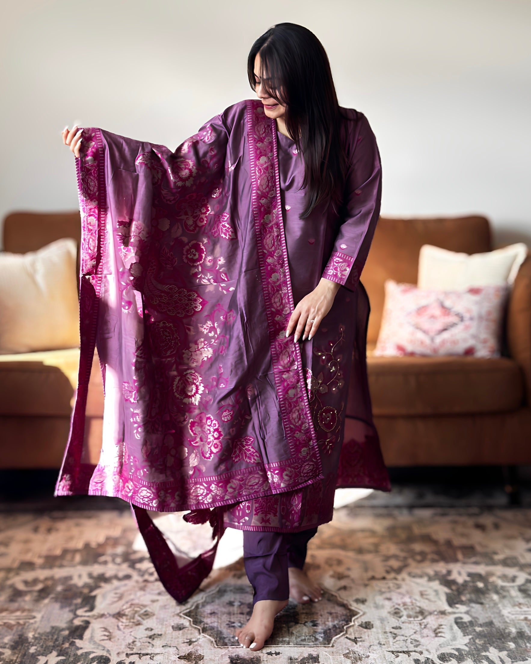 Woman in a purple traditional outfit holding a matching dupatta in a living room.