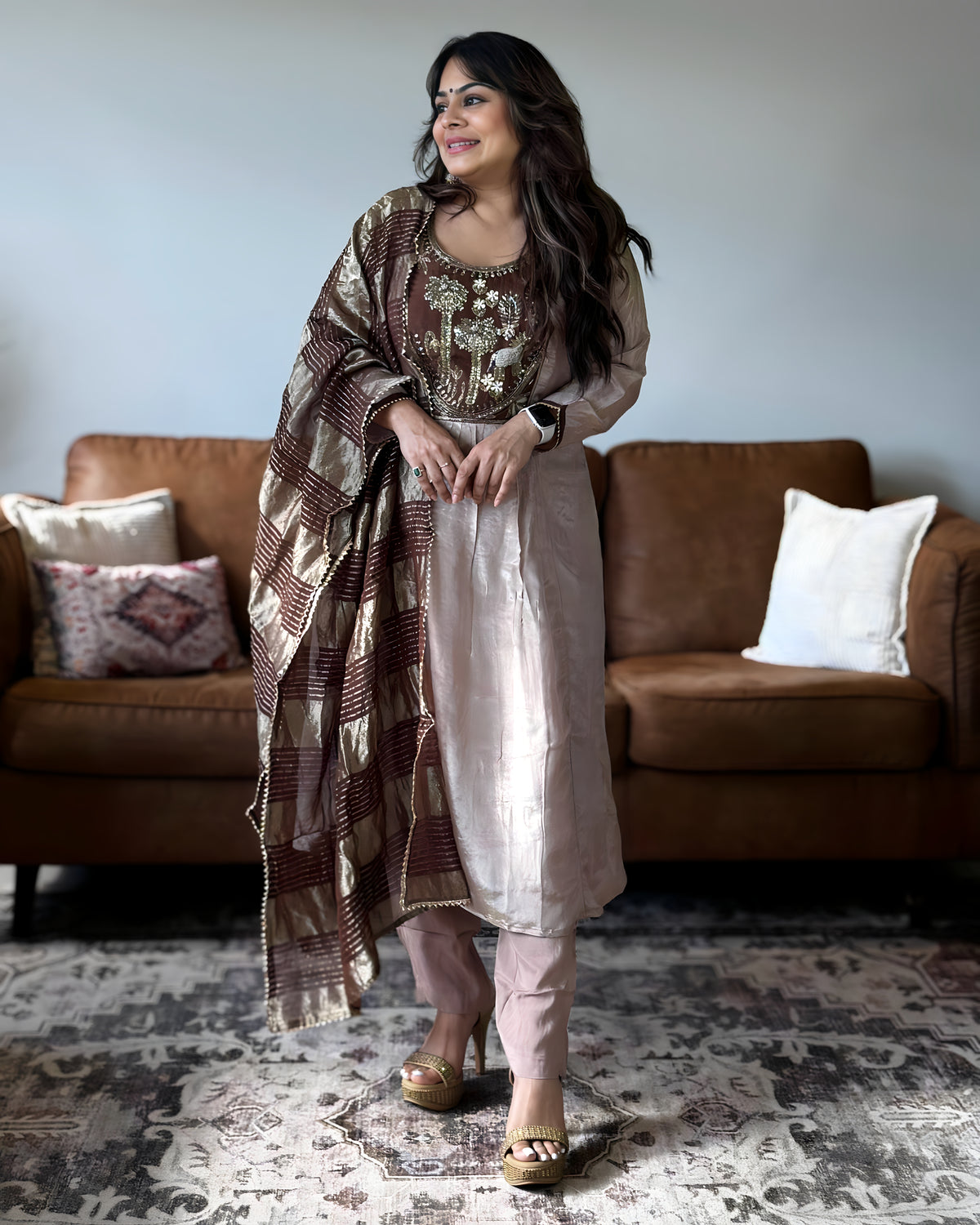 A woman models a traditional South Asian outfit consisting of a beige kurta with intricate brown and gold embroidery, matching trousers, and a patterned brown and gold dupatta, standing in a living room in front of a brown leather sofa.