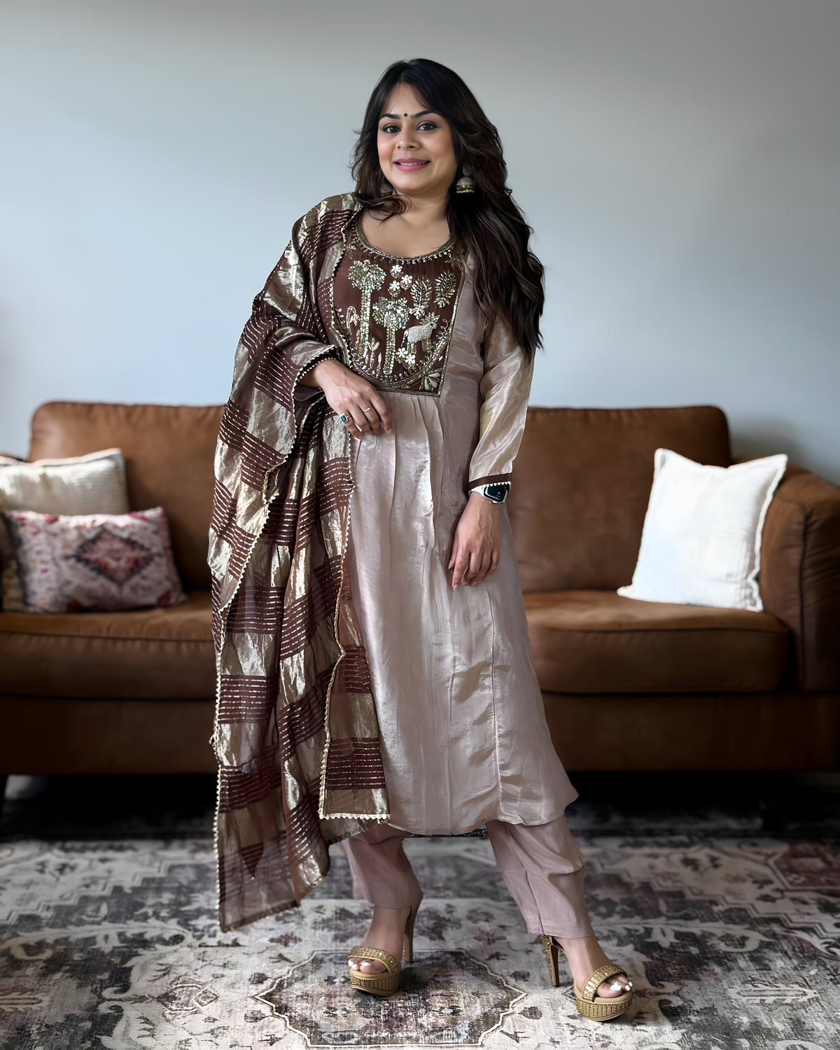 A woman models a traditional Indian salwar kameez suit set in a living room setting. The outfit features a light beige satin kurta with intricate gold embroidery on the chest, matching beige trousers, and a brown and beige patterned dupatta draped over her shoulder.