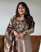 A woman wearing a traditional Indian outfit with a brown and gold embroidered kurta and a matching patterned dupatta, standing in front of a plain light gray wall.