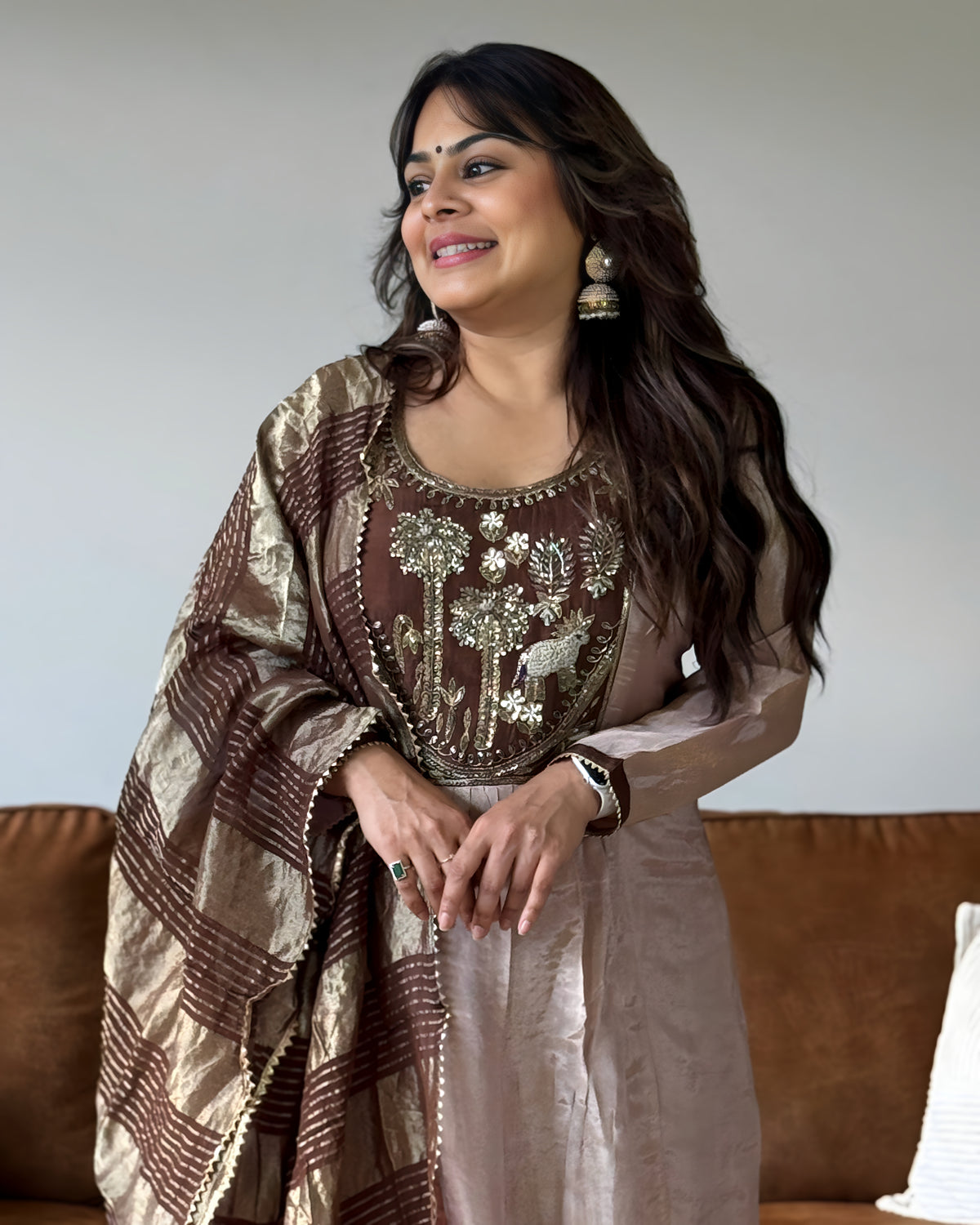 A woman wearing a traditional Indian outfit with a brown and beige patterned dupatta, a brown embroidered blouse, and a light beige kurta, standing in front of a brown sofa.