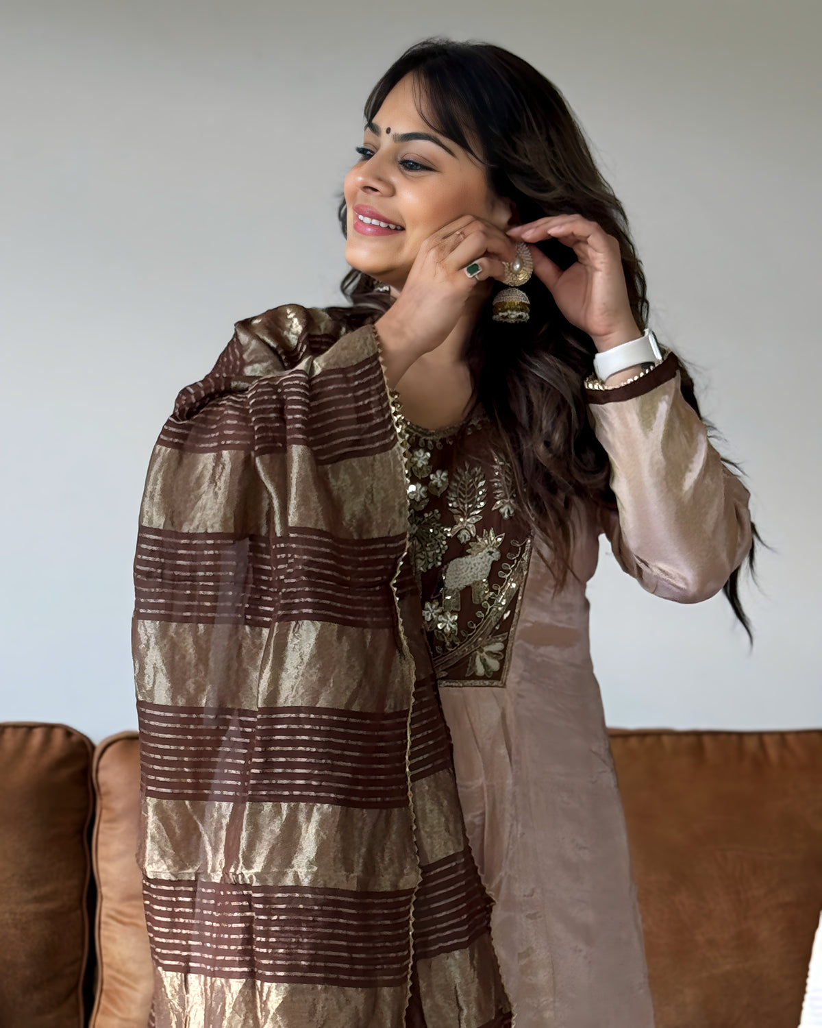 A woman wearing a traditional South Asian outfit with a metallic gold and brown striped dupatta draped over her shoulders, a beige embroidered kurta, and gold earrings, standing in front of a brown sofa.