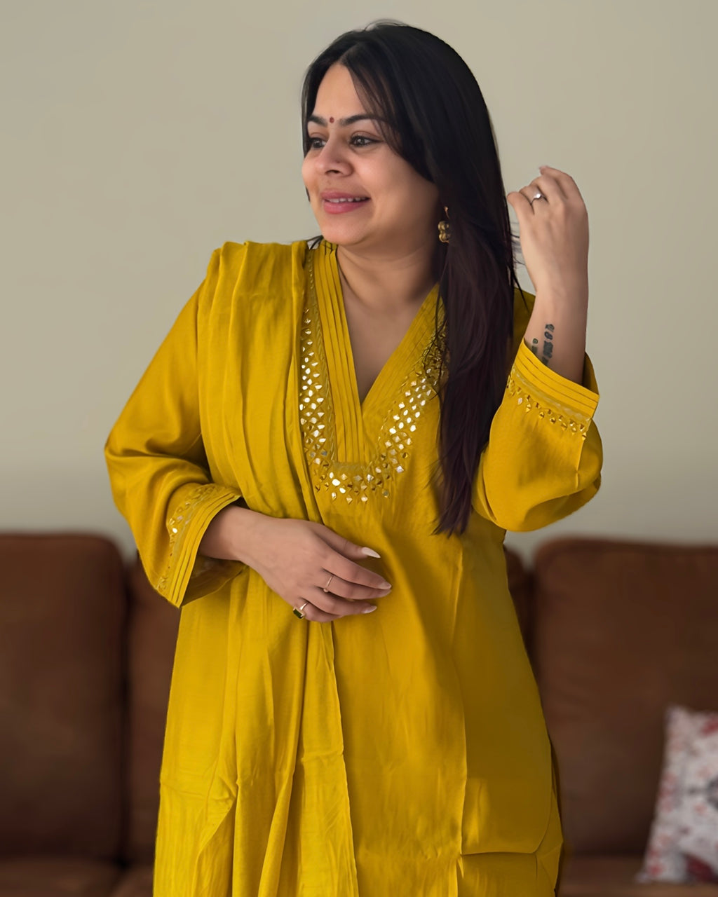 Woman wearing a yellow dress with intricate designs, standing in a room with a brown couch and patterned pillow.