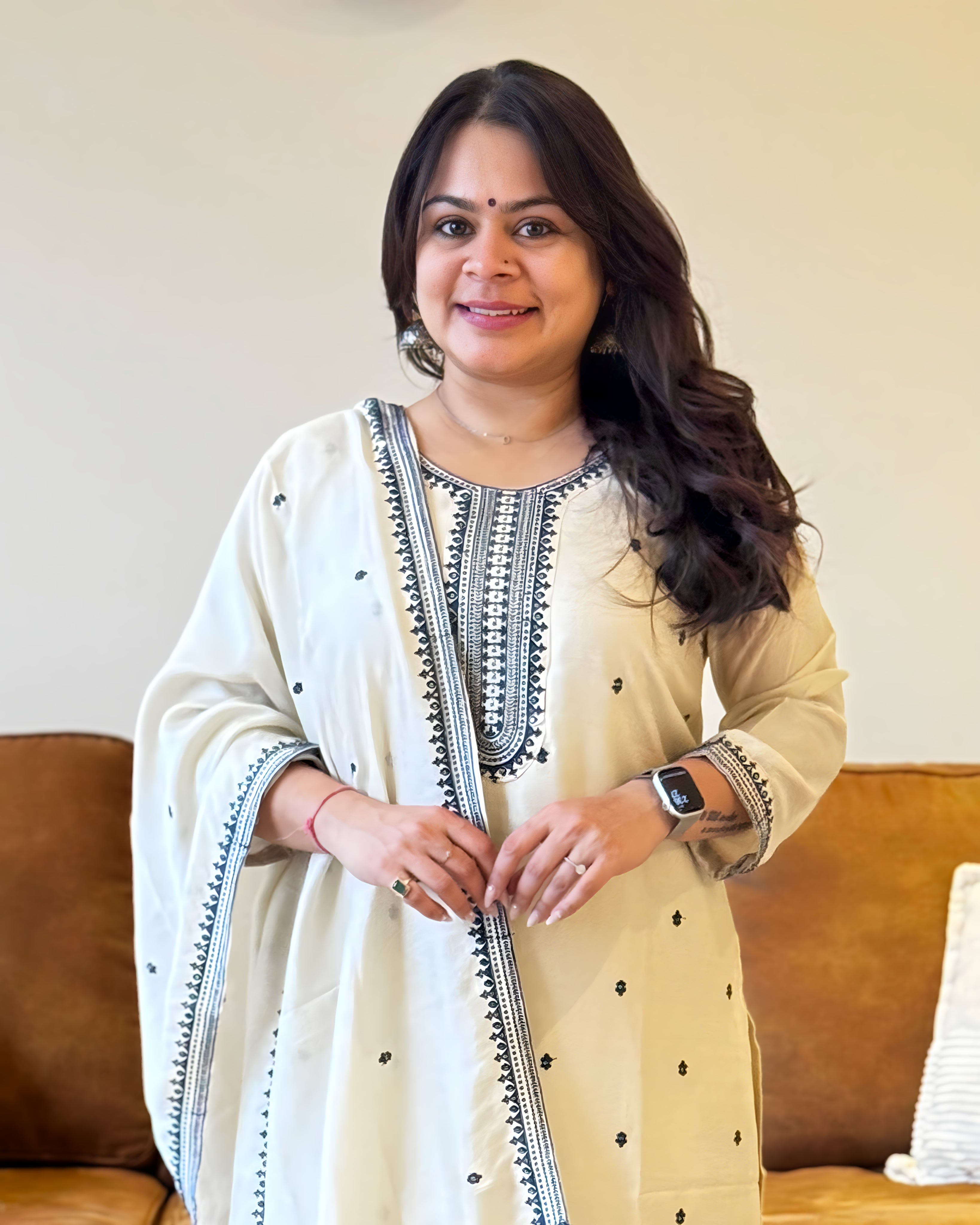 Woman wearing a cream-colored traditional outfit with black embroidery in a room.