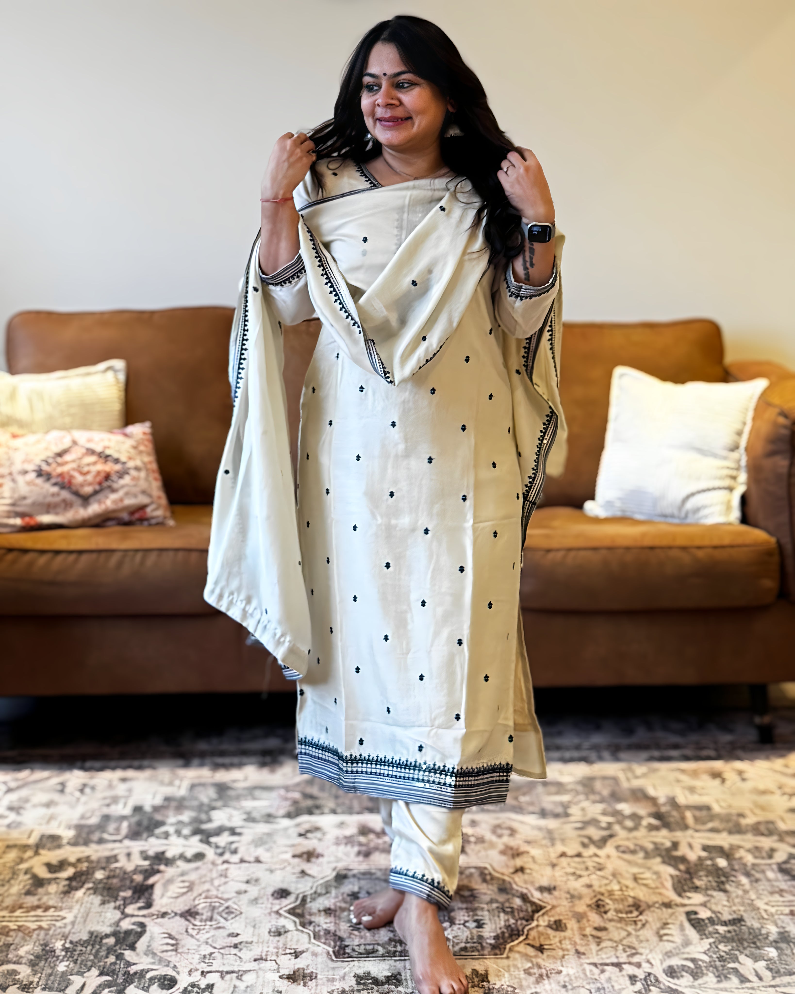 Woman in a traditional outfit standing in a living room with a brown sofa and patterned rug.