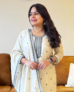 Woman wearing a cream-colored traditional outfit with black embroidery sitting on a brown couch.