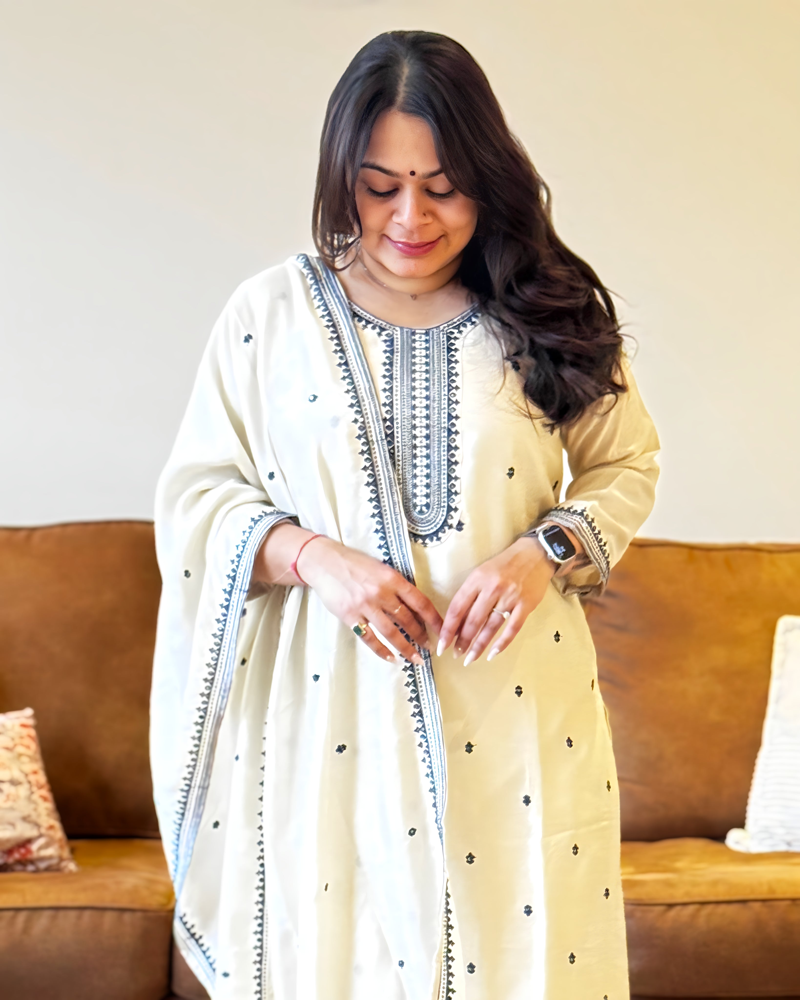 Woman wearing a cream-colored traditional outfit with black embroidery sitting on a brown couch.