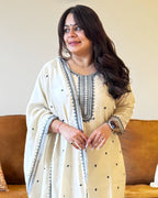 Woman wearing a beige and white traditional outfit with intricate patterns, standing against a neutral background.