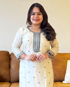 Woman wearing a white traditional outfit with black embroidery in a living room setting.