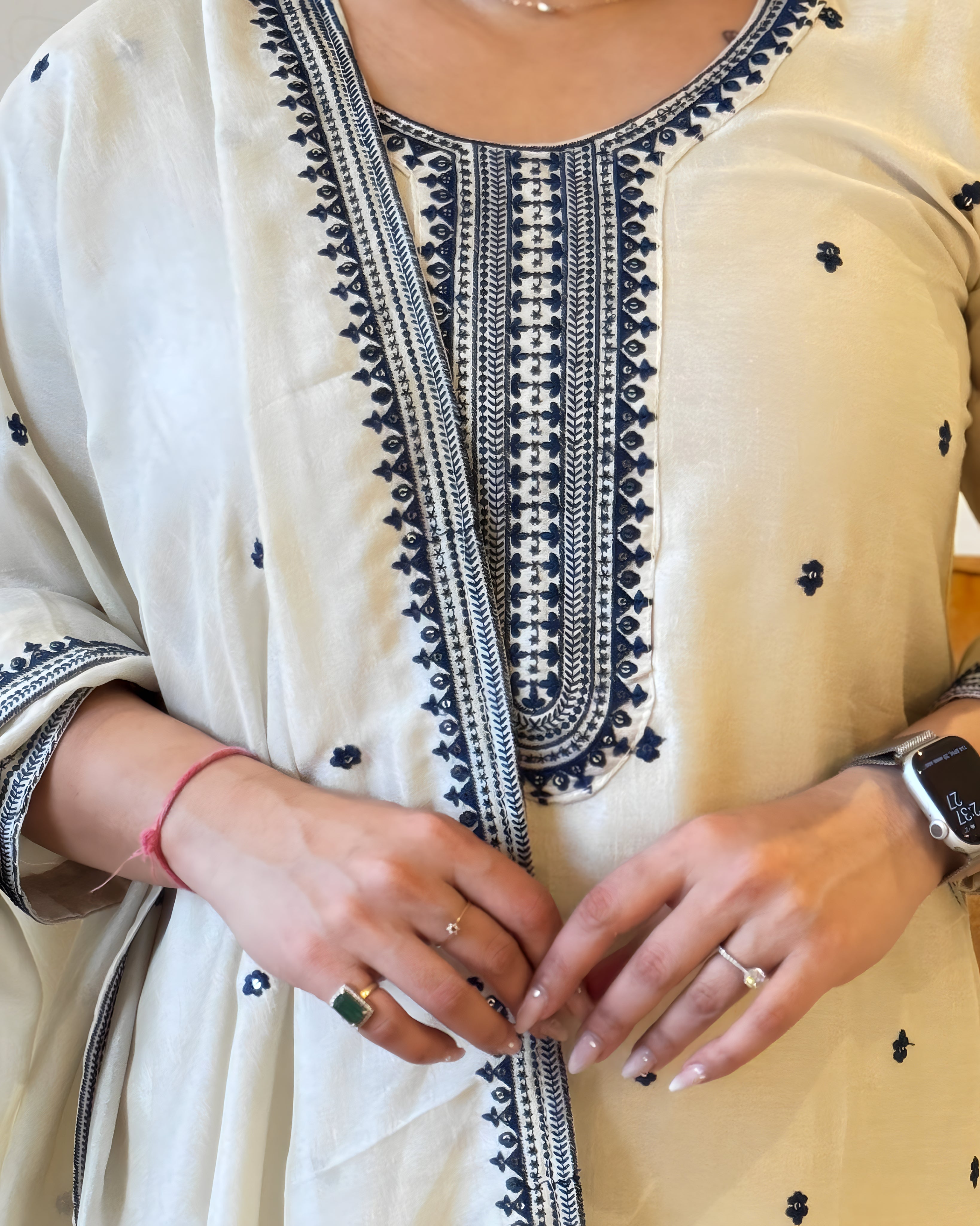 Close-up of a person wearing a cream-colored outfit with black embroidery, focusing on the neckline and hands.