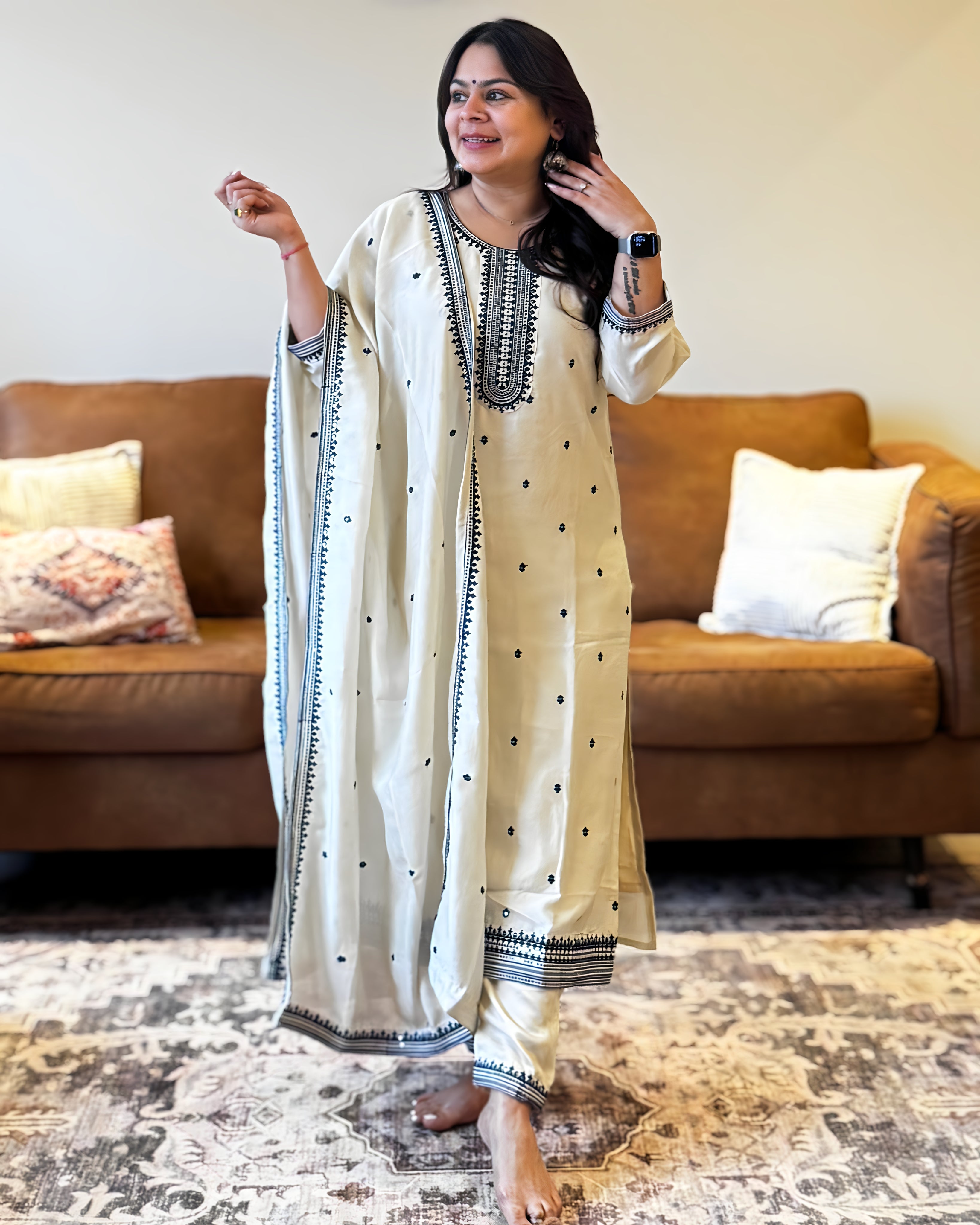 Woman wearing a cream-colored traditional outfit with black patterns in a living room.