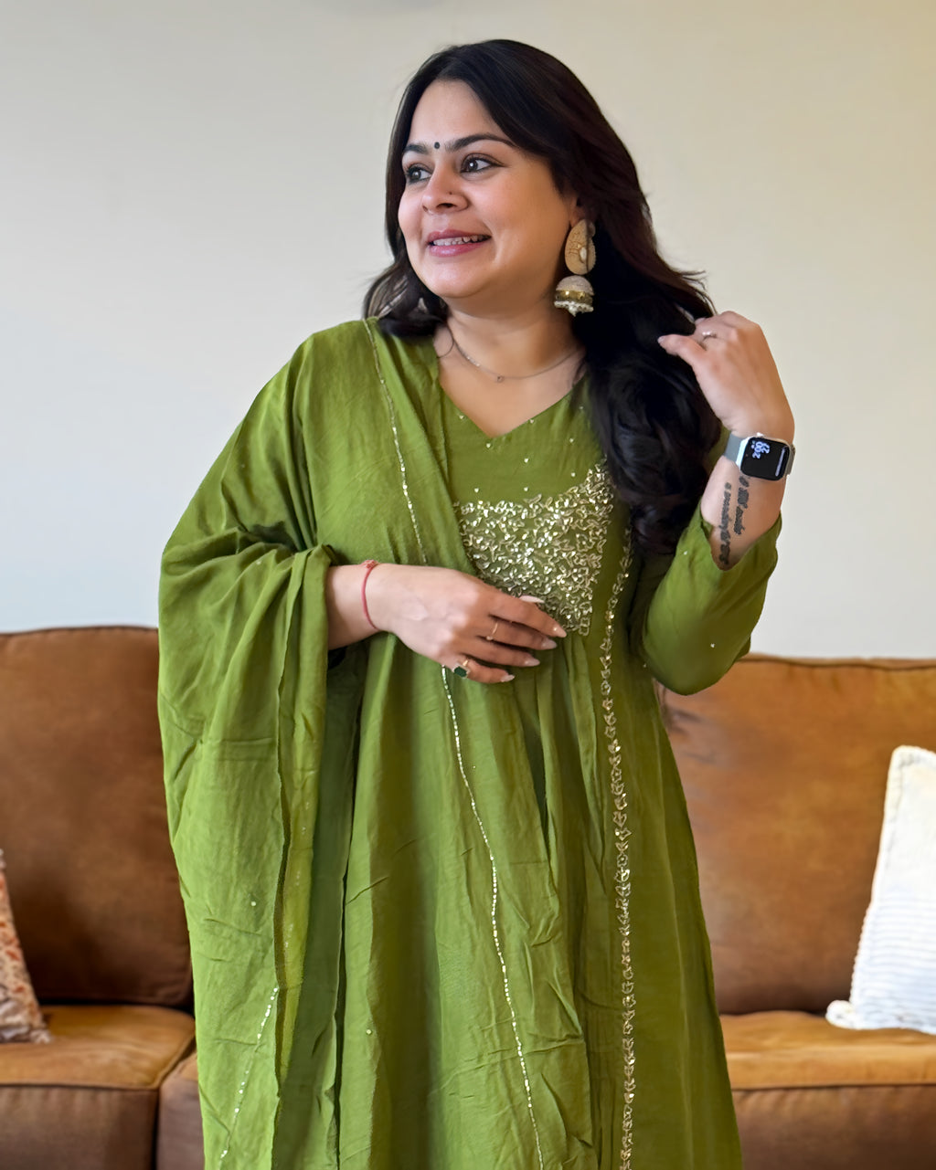 Woman wearing a green traditional outfit with a sequined top, standing in a room with a brown couch.