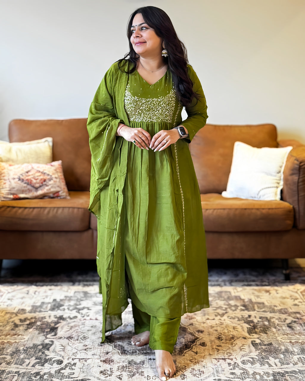 Woman in a green traditional outfit standing in a living room with a brown sofa and patterned rug.