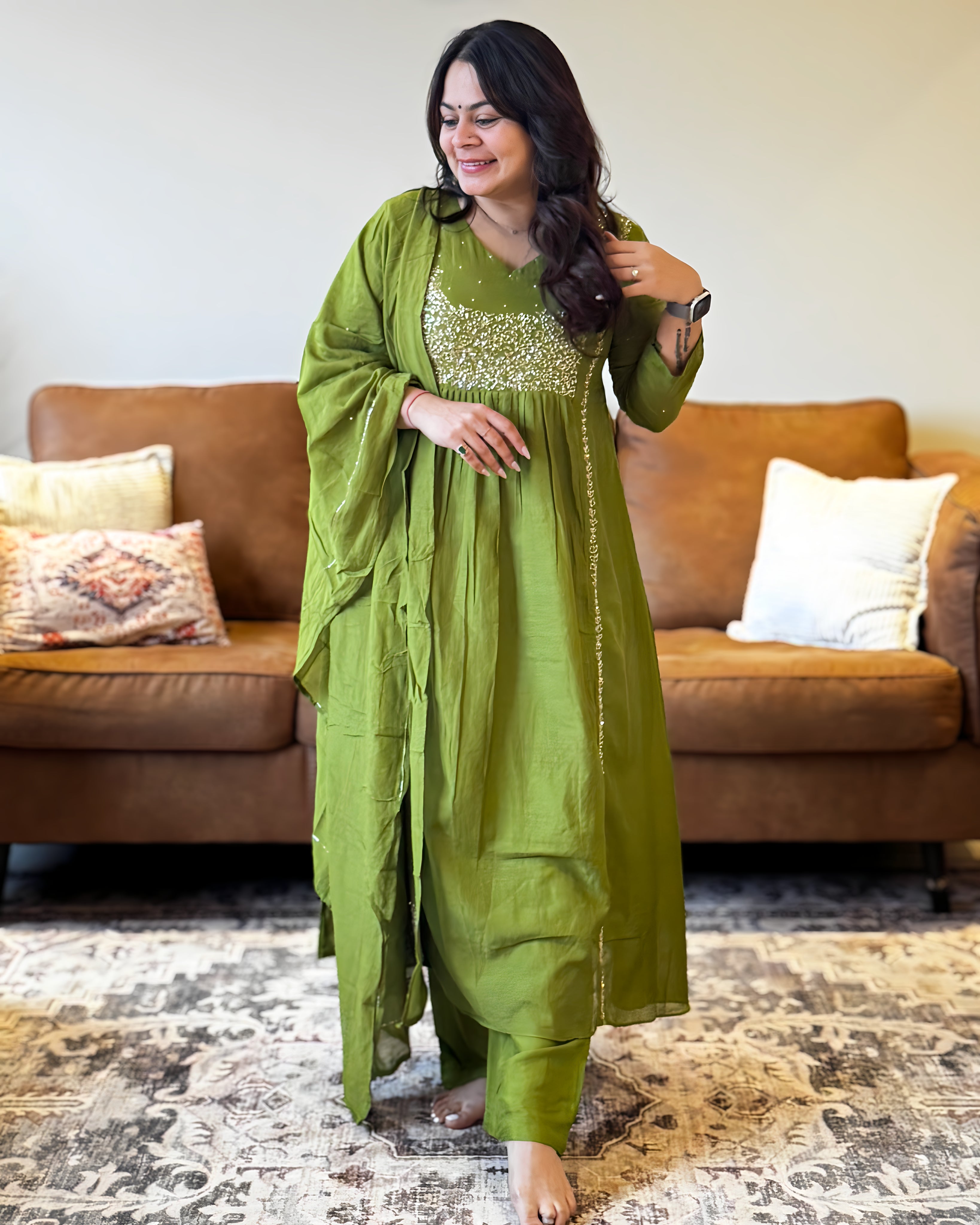 Woman in a green traditional outfit standing in a living room with a brown sofa and patterned rug.