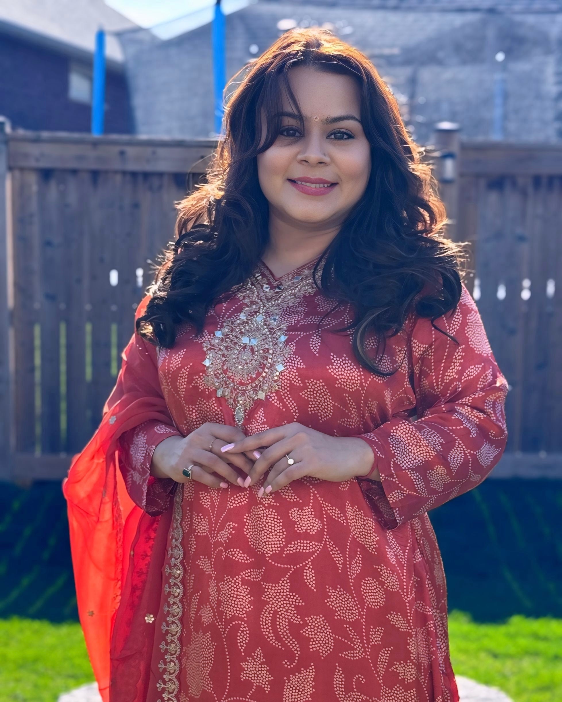 Woman wearing a red patterned dress standing outdoors with a wooden fence and grass in the background.