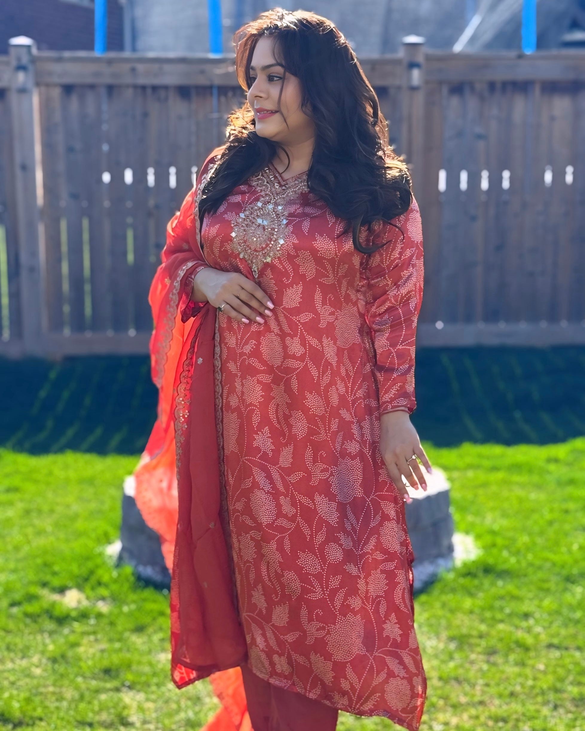 Woman in a red floral dress standing outdoors on grass with a wooden fence in the background