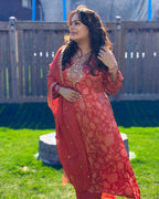 Woman in a red traditional outfit standing outdoors on grass with a wooden fence in the background.