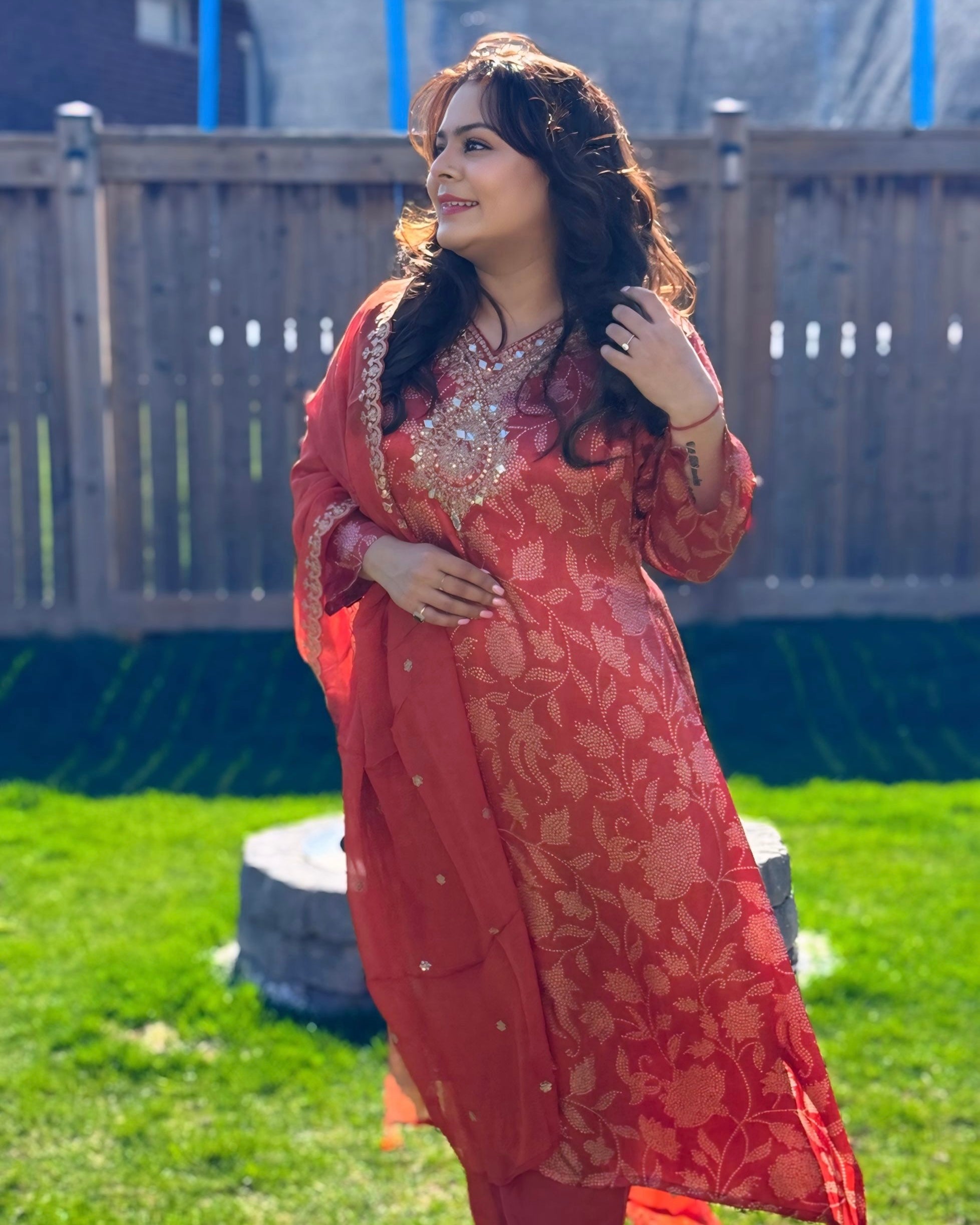 Woman in a red traditional outfit standing outdoors on grass with a wooden fence in the background.