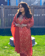 Woman in a red traditional outfit standing outdoors on grass with a wooden fence in the background.