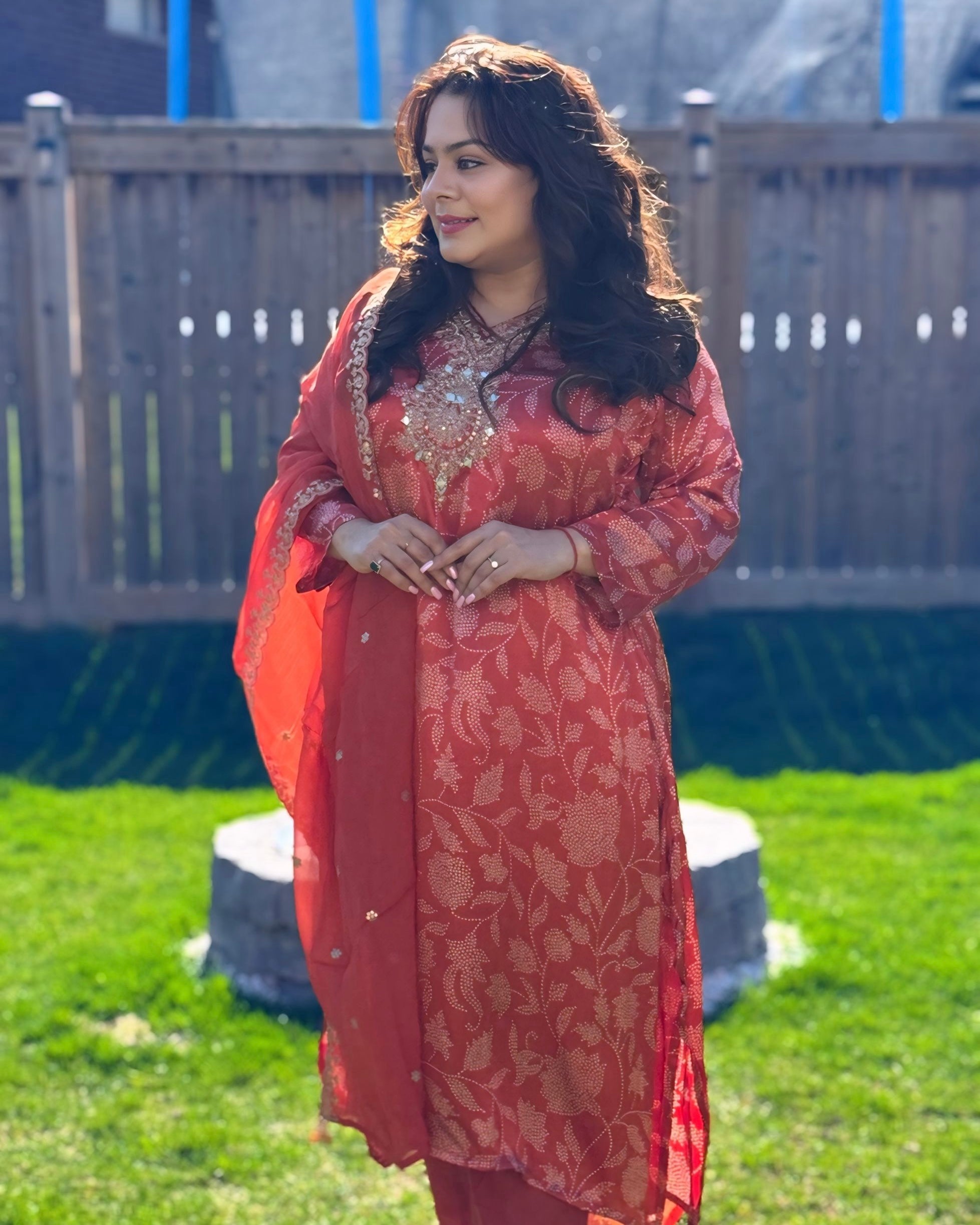 Woman in a red traditional outfit standing outdoors on grass with a wooden fence in the background.