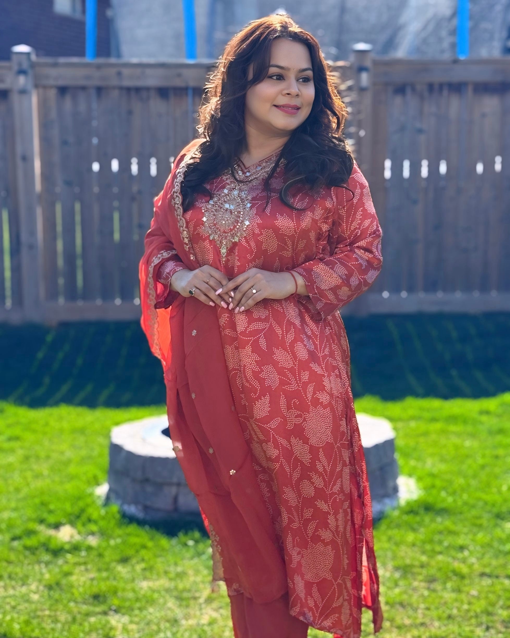 Woman in a red traditional outfit standing outdoors on grass with a wooden fence in the background.