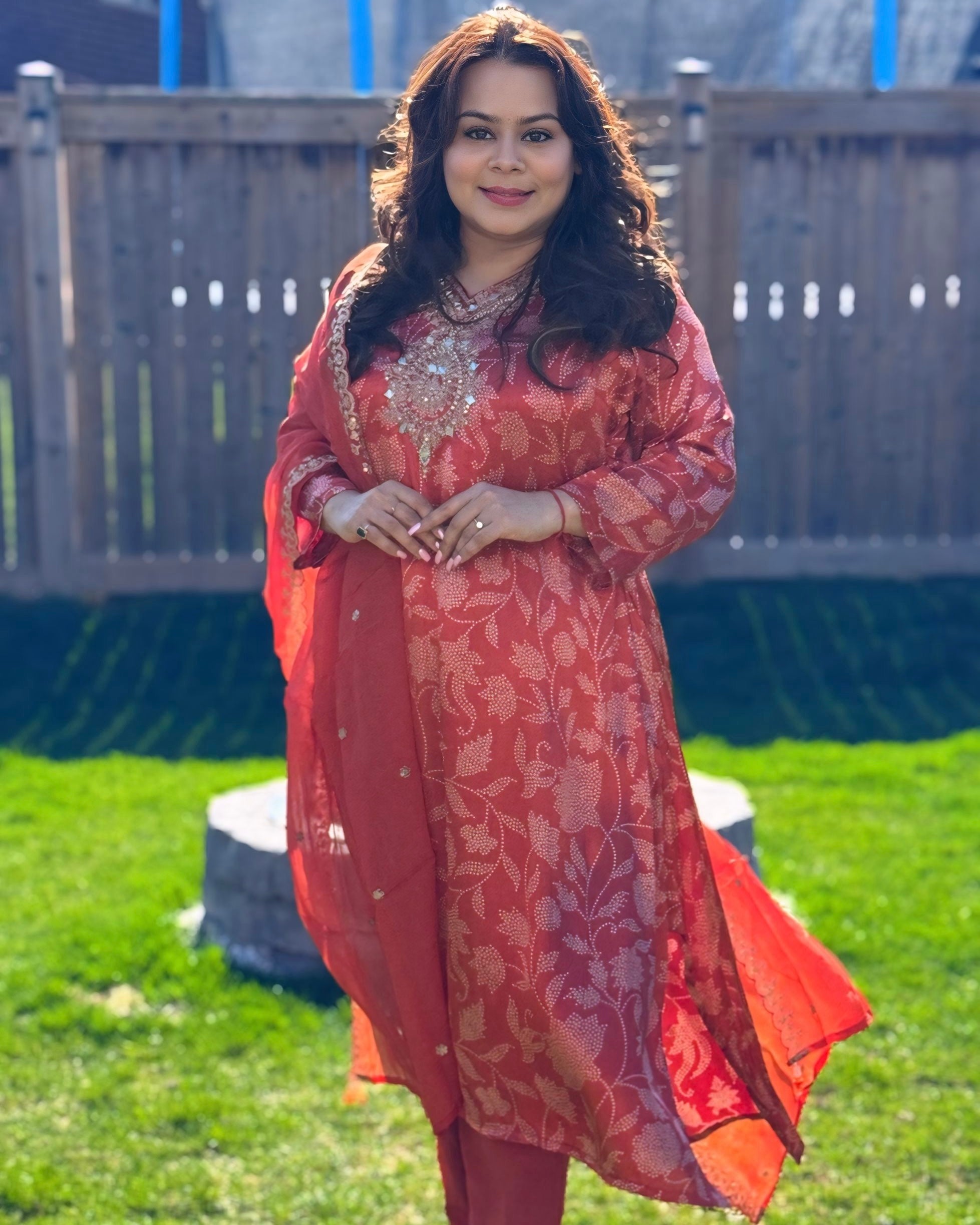 Woman in a red traditional outfit standing outdoors on grass with a wooden fence in the background.