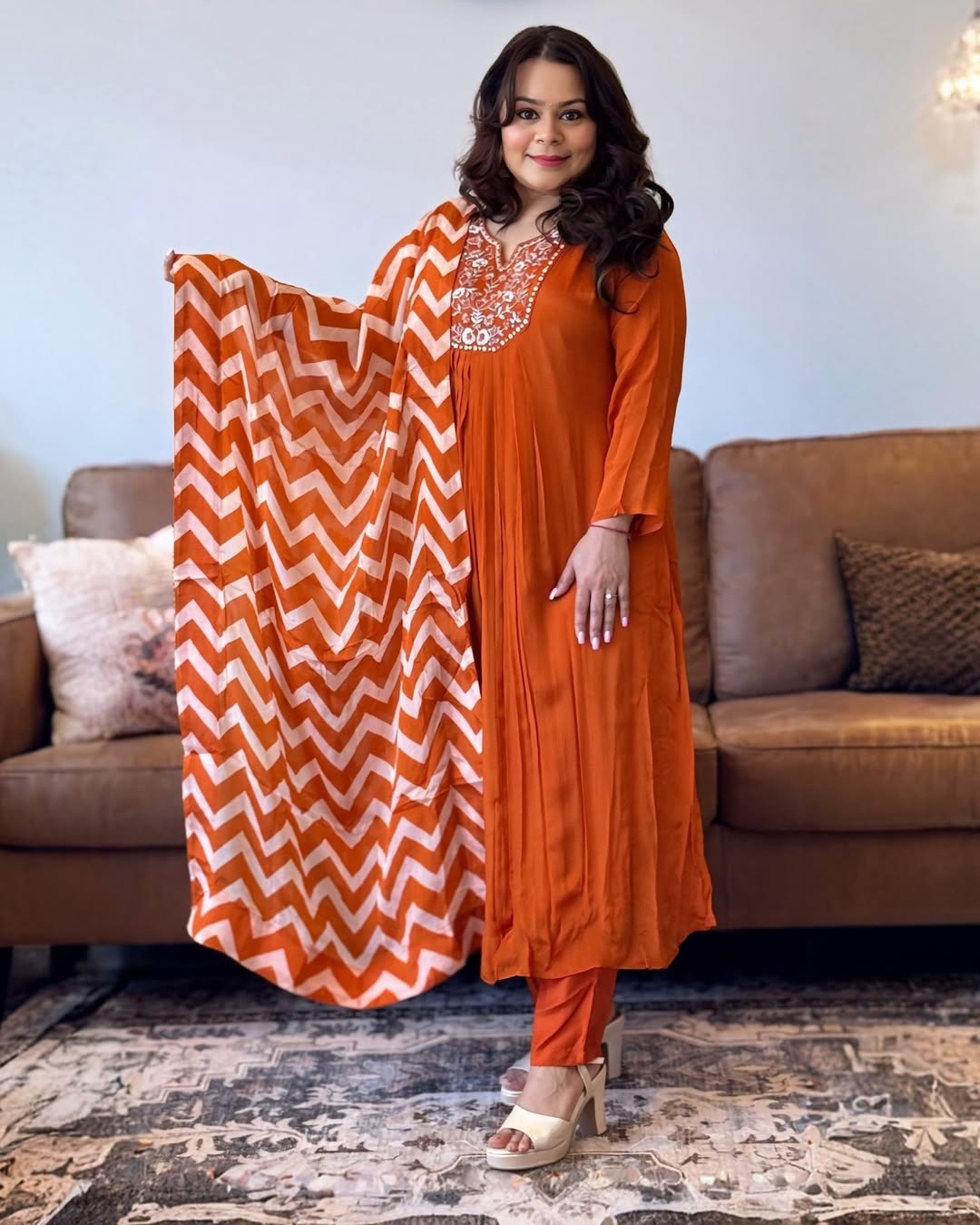 Woman in an orange traditional outfit with a white zigzag pattern, standing in a living room.